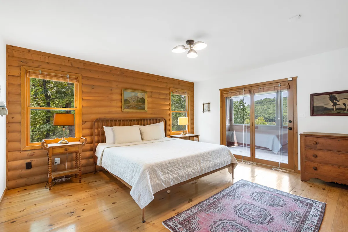 This image shows a calm and tranquil bedroom with wooden walls, a cozy bed, a soft rug, and large windows that let in abundant natural light. The inviting furniture and serene atmosphere make it an ideal private retreat.