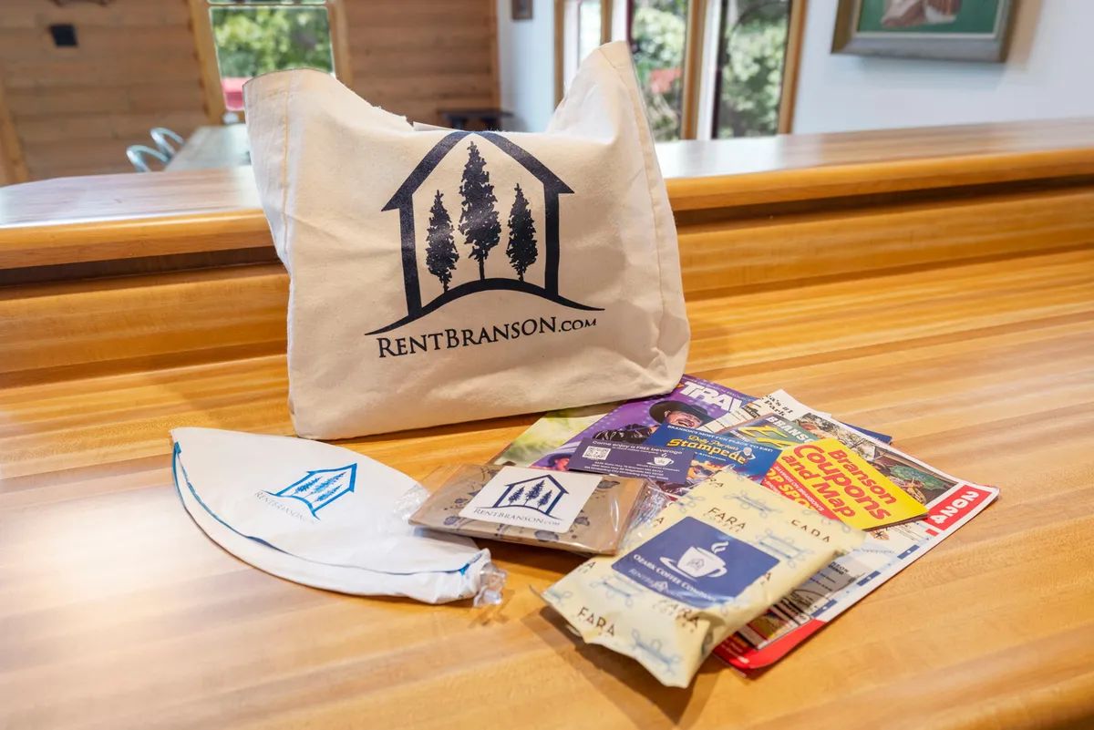 A canvas tote bag bearing the property logo sits on a countertop, surrounded by an assortment of brochures, maps for local attractions, and small snack packets. This welcoming arrangement signals thoughtful preparation and provides resources for exploring the area.