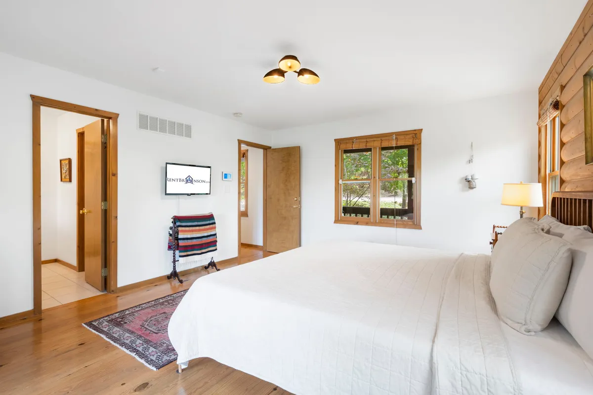 A serene bedroom furnished with a comfortable bed, a large window providing plenty of natural light, a TV, a soft rug, and a classic wooden door. The overall feeling is calm and relaxing—perfect for unwinding after a day of adventure.