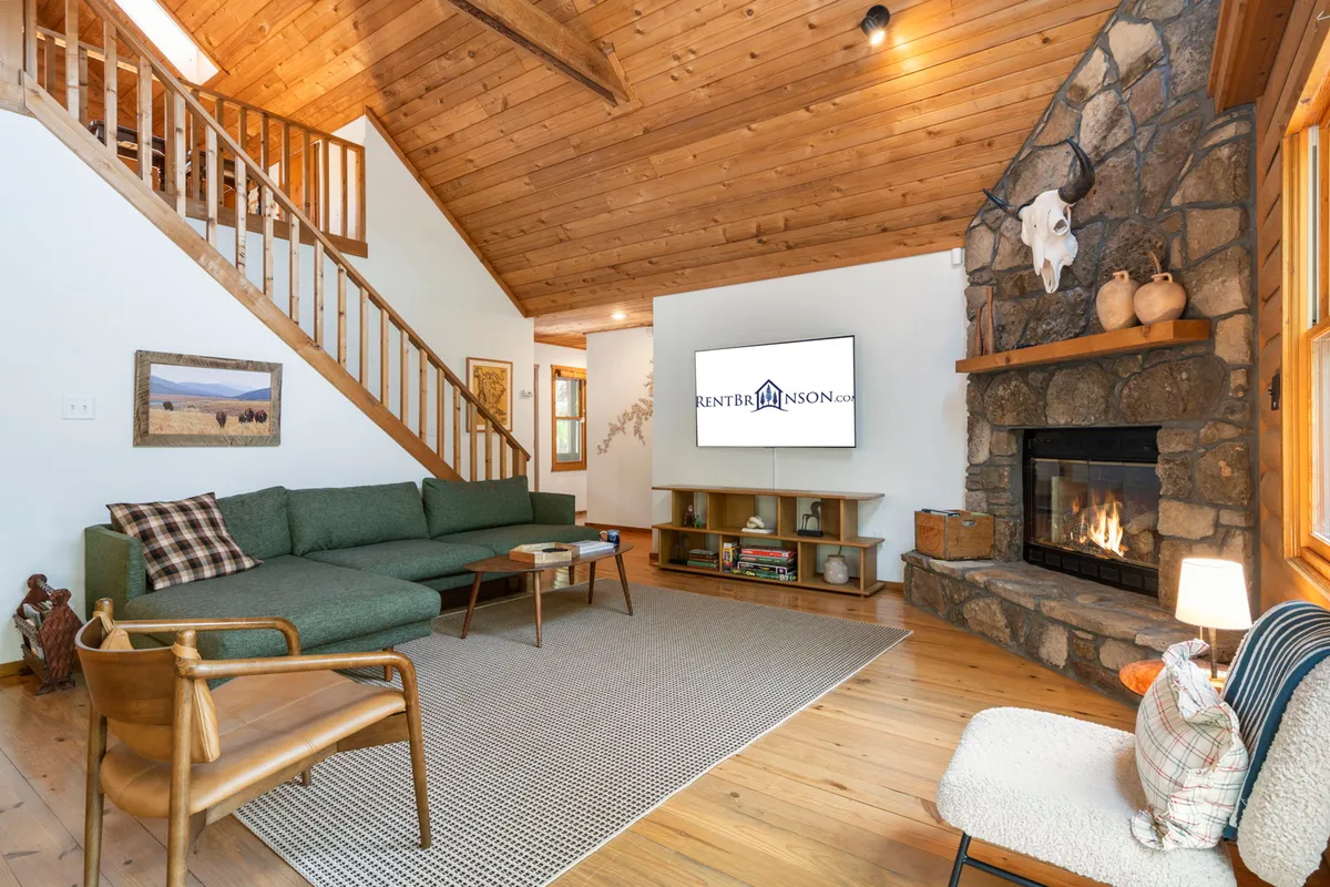 A cozy living room scene centered around a stone fireplace, with a large green sofa, wooden staircase, and wall-mounted television. The setup encourages relaxation and socializing, perfect for movie nights or board games by the fire.