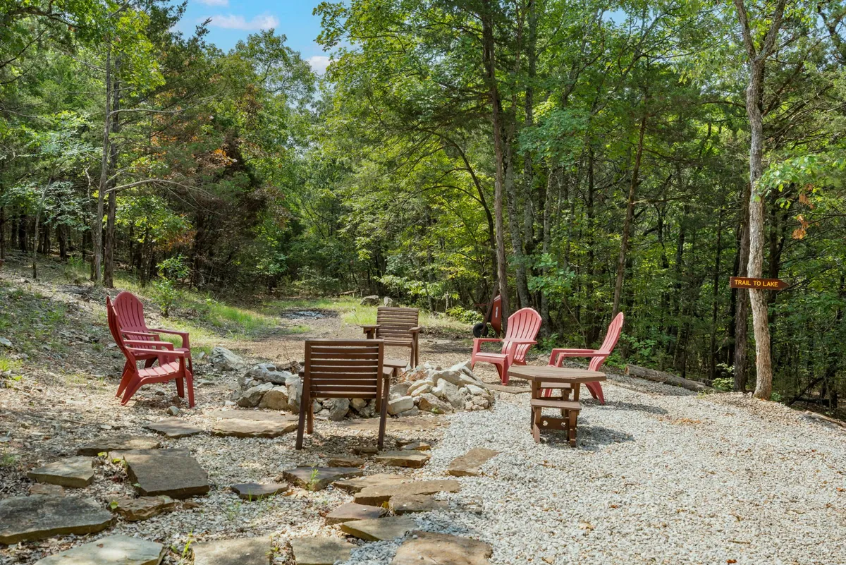 A cozy fire pit encircled by red Adirondack chairs, nestled among trees and connected by a gravel pathway. The space is set up for communal gatherings, marshmallow roasts, and evening stargazing.