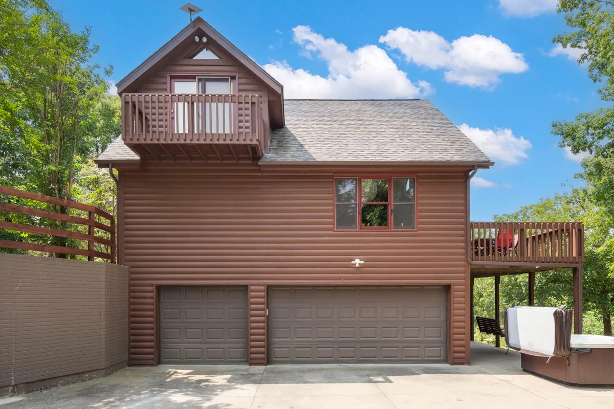A broad wooden balcony overlooks green trees, with ample room for relaxing or dining outdoors. The cozy exterior includes large windows and a welcoming vibe, inviting guests to sit outside and enjoy the tranquil setting.