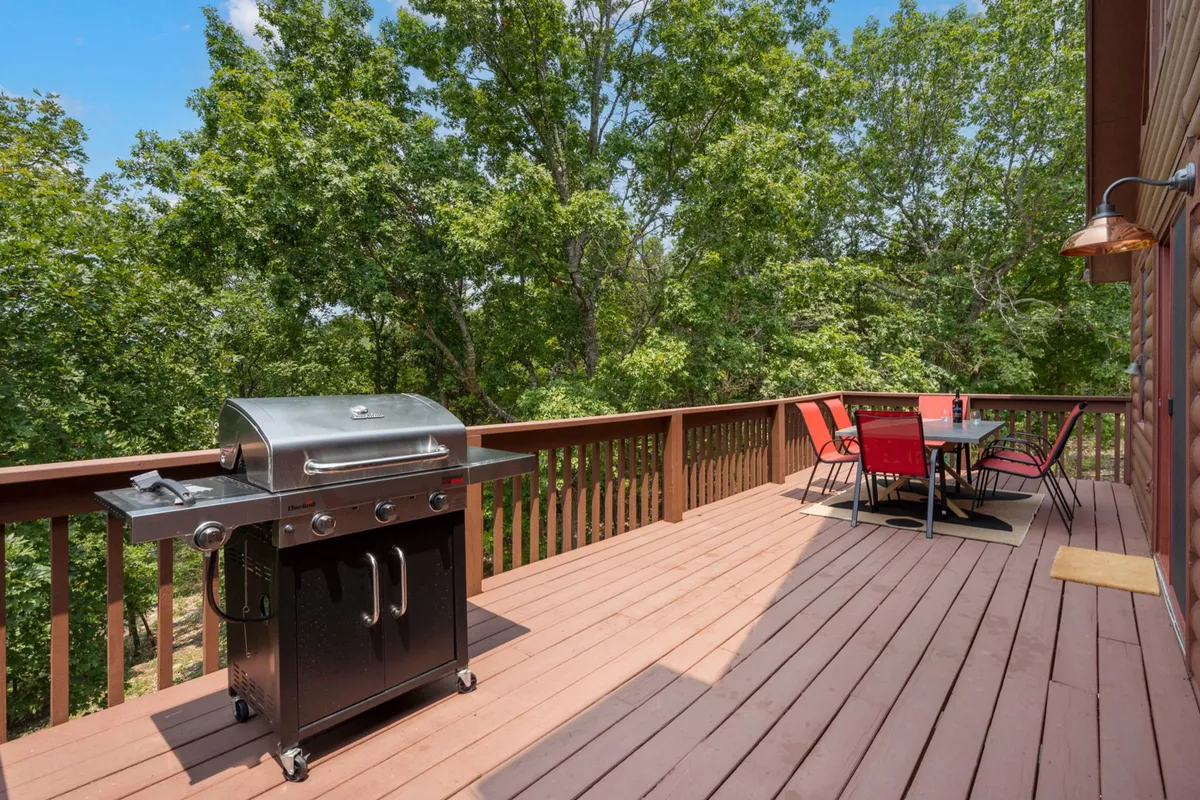 This image features a grill and outdoor dining table set on a spacious deck, surrounded by lush trees. The relaxed vibe is perfect for gatherings, with ample space for alfresco meals and sunset enjoyment.