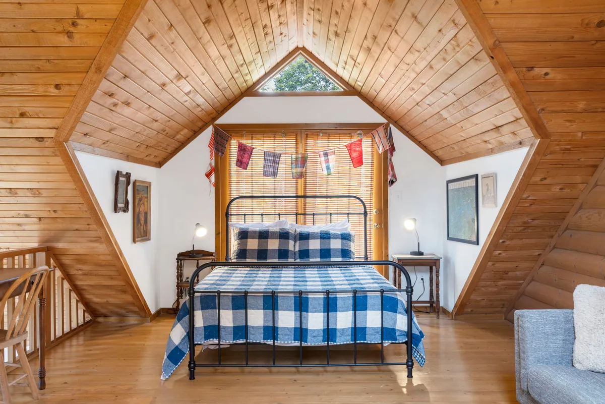 A warm and welcoming bedroom featuring a bed with a plaid coverlet, decorative flags above, wooden beams and walls, and charming wall art. The space exudes a sense of rustic coziness and personal touch, making it feel like a true retreat.