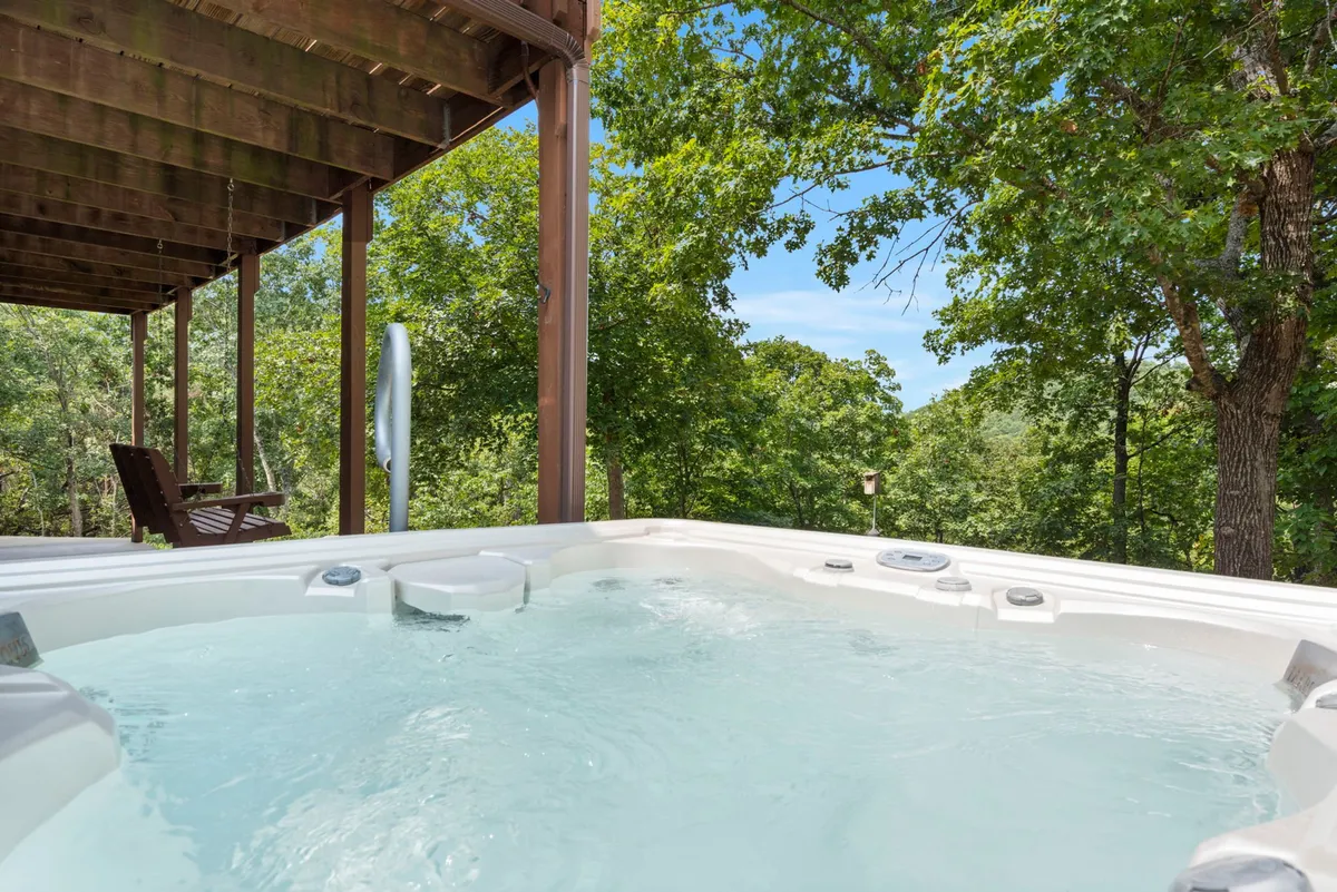 A tranquil exterior view of a deck area with a private hot tub set amid lush greenery, ideal for starlit soaks. The surrounding treetops and open sky offer an inviting setting for relaxation and connection to nature.