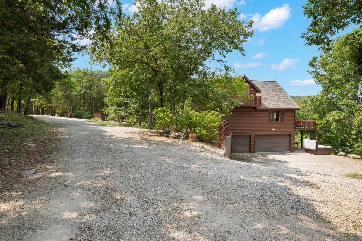 This image showcases the exterior of Buffalo Bluff, with a charming wooden house set among tall trees and a gravel driveway. The lush greenery hints at the natural beauty and year-round appeal of the Ozarks, emphasizing the property's connection to outdoor experiences.