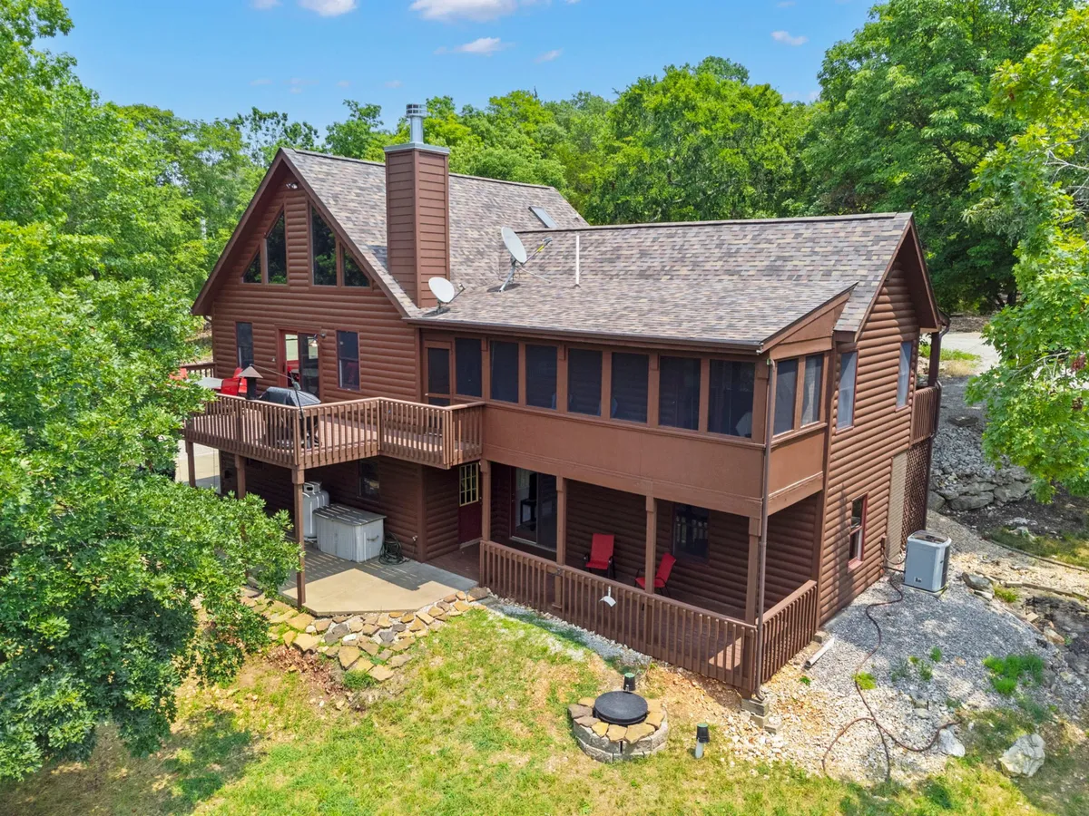 A broad exterior view of the large wooden house set amid lush greenery, with inviting red chairs on the deck and a thoughtfully arranged fire pit area. The scene is bathed in cozy evening light, evoking a sense of togetherness and outdoor enjoyment.