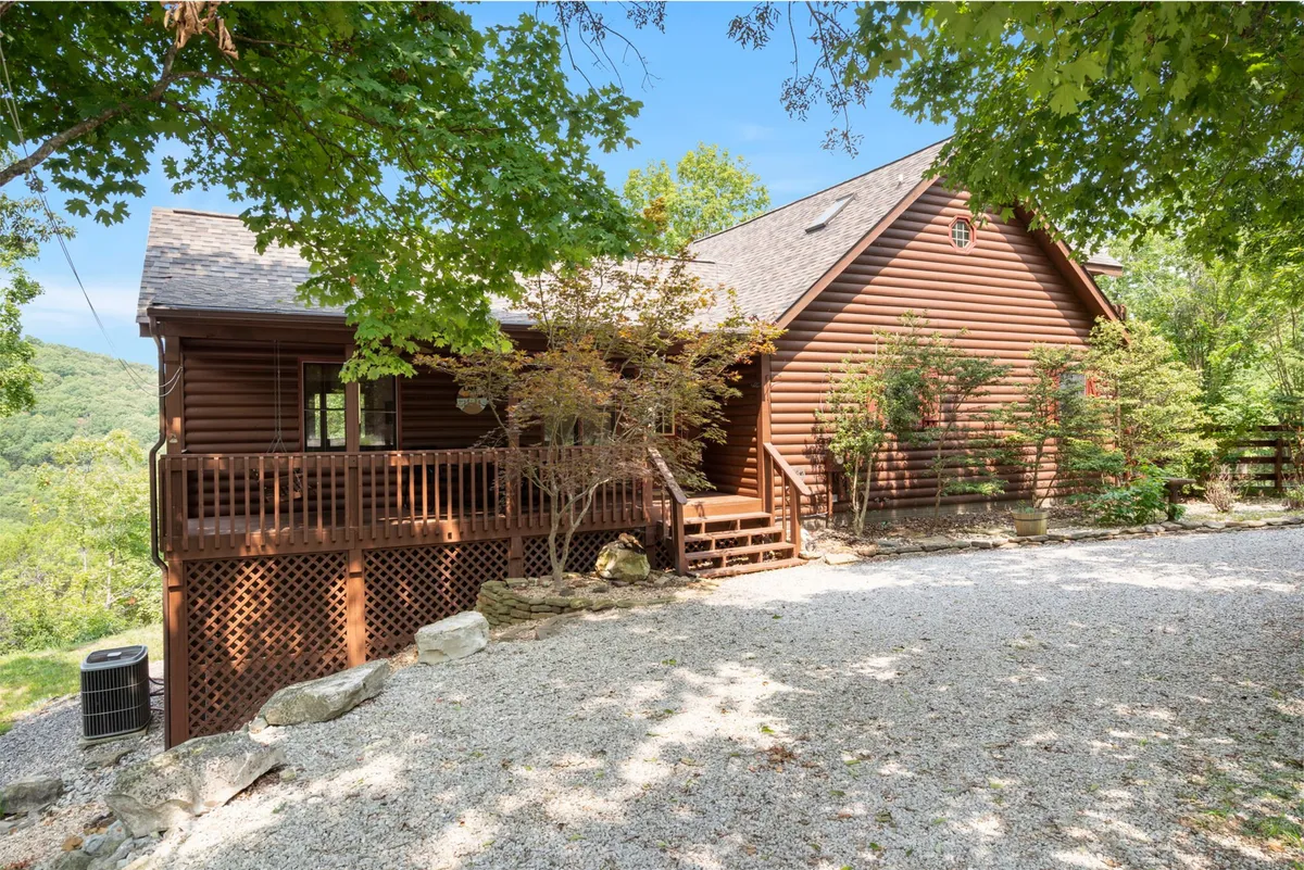 The image features the log cabin structure with a front porch accented by railings and surrounded by lush greenery. The setting is tranquil, and the porch looks inviting for both people and pets to relax or watch the sunset after a day of adventures.