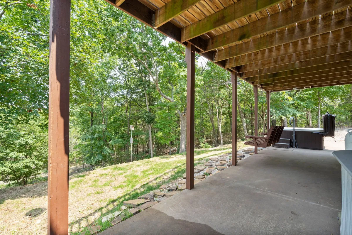 A serene patio area beneath sturdy wooden beams, surrounded by greenery and shaded for comfort. This outdoor space is ideal for relaxing or entertaining, with ample natural beauty for both people and pets to enjoy.