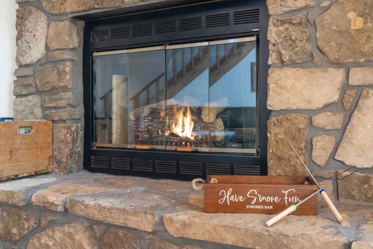A cozy interior scene featuring a fireplace, a 's’mores bar' sign, and stacked firewood. The setting invites family gatherings around the fire and is symbolic of warm winter nights, fireside chats, and making memories regardless of the season.