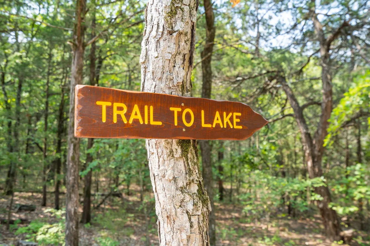 A naturally framed outdoor pathway marked by a wooden sign, winding through a wooded area. The scene suggests adventure and access, inviting guests to explore the private path leading to the lake shoreline.