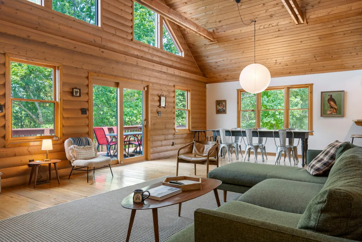 A bright, spacious living area featuring large windows that showcase lush greenery outside. The room is anchored by a comfortable sectional sofa and a stylish light fixture. The adjoining dining area, with modern metal chairs, is visible, creating an inviting and open atmosphere.