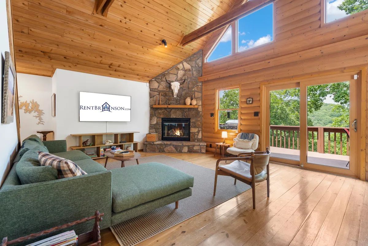 The image captures a cozy living area featuring a striking stone fireplace, plush green sofa, and expansive windows that fill the room with natural light and showcase tranquil wooded views. A wooden deck is visible through the windows, hinting at seamless indoor-outdoor living and the promise of serene sunset moments. The warm tones and inviting layout evoke comfort and relaxation.