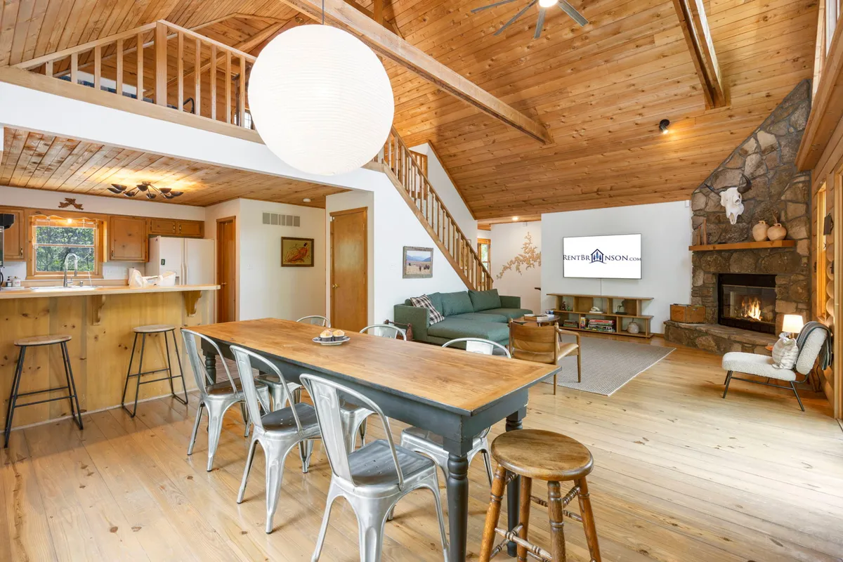 This image presents a spacious open-concept main level, featuring a large dining table, a visible fireplace, ample living area seating, and a kitchen bar. The blend of wood tones and warm lighting embodies the cabin’s welcoming ambiance. Guests can easily imagine gathering here for shared meals or relaxing by the fire.