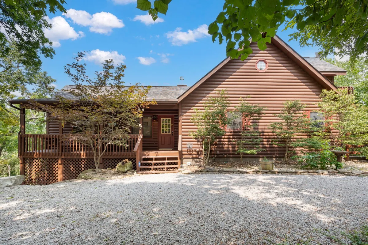A cozy log cabin with a welcoming front porch, surrounded by well-maintained landscaping and a gravel driveway. The porch is set up for relaxation and socializing, offering a perfect spot to watch sunsets or enjoy the fresh Ozark air with your pet after a day of exploring.
