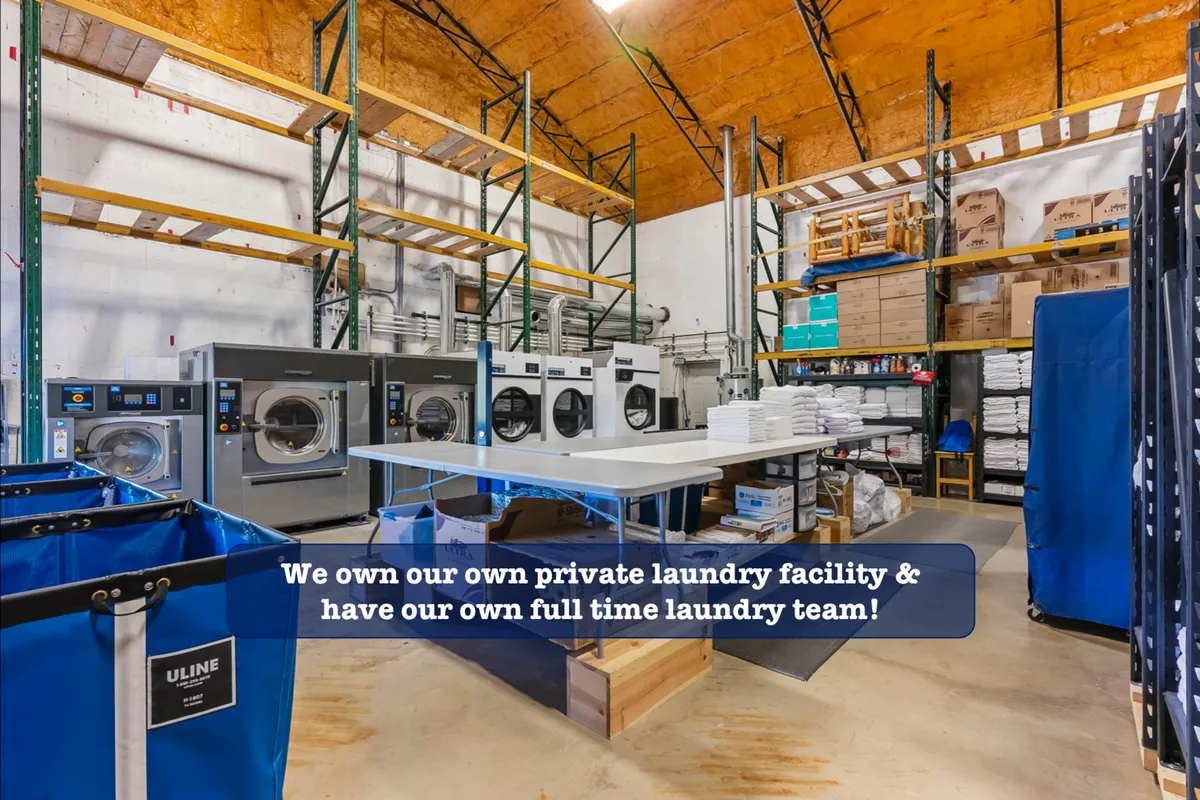 This image shows a commercial laundry facility with large industrial washing machines, a folding table, and laundry bins. The scene conveys a sense of professionalism and cleanliness, emphasizing the behind-the-scenes care that goes into maintaining linens and towels.